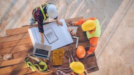 3 construction workers look over a table with blueprints, work equipment, and a laptop